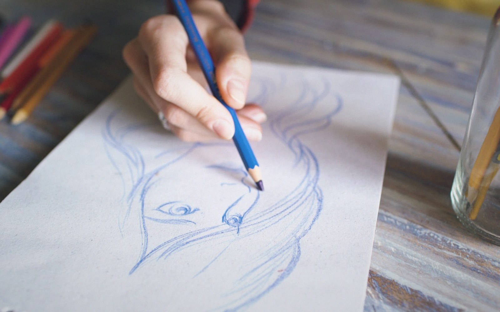 A hand sketches a face and flowing hair using a blue pencil on paper