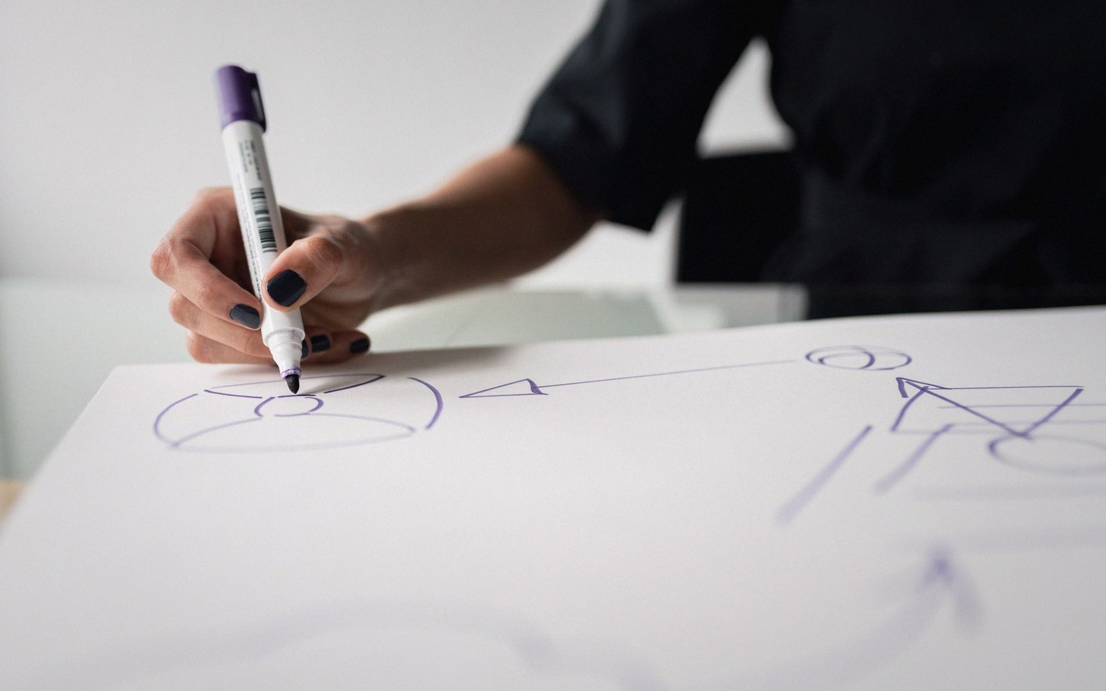 A hand with dark nail polish sketches diagram shapes with a purple marker on paper