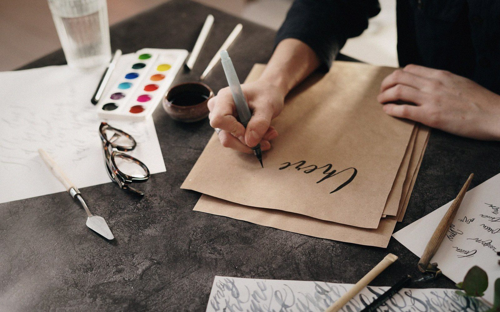 A hand practices calligraphy with a dip pen on kraft paper beside watercolors and glasses